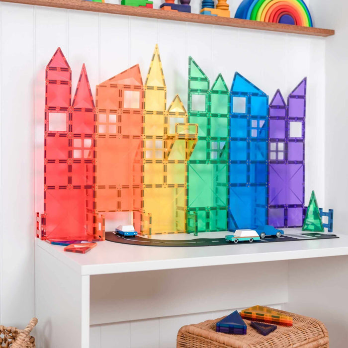 Rainbow magnetic tiles built into tall skyscraper towers with toy cars on a black road atop a white desk.
