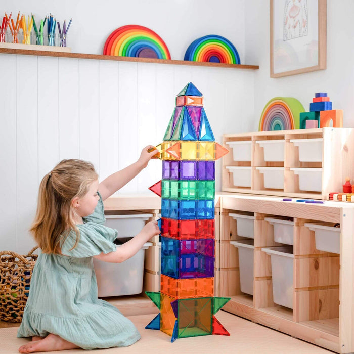 Child building a tall rainbow rocket using colorful square and triangle magnetic tiles in a bright playroom.