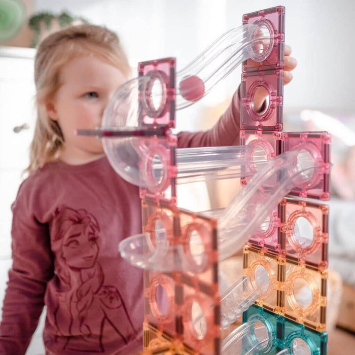 Young girl building a tall pastel magnetic tile tower with clear chutes and a pink rolling ball.