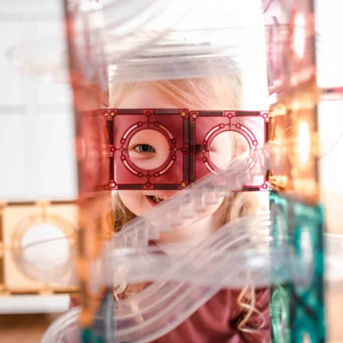 Child smiles through a pink translucent magnetic tile hole, part of a ball run set with clear plastic tracks.