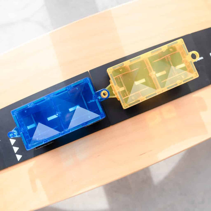 Transparent blue and yellow magnetic car bases connected on a black roadway track over a light wood table.