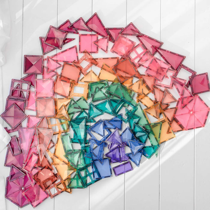 Pastel magnetic tiles in various geometric shapes arranged in a large rainbow swirl on a white wooden surface.