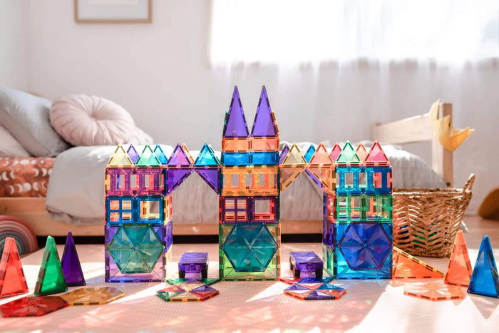 Rainbow magnetic tile bridge and towers with geometric windows and car bases on a light pink playroom rug.