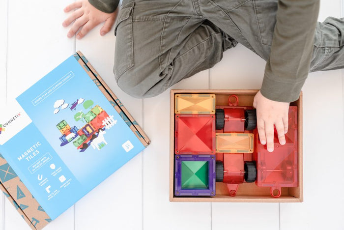 Child reaching into a box of colorful magnetic tiles and red wheeled bases next to a blue Connetix box lid.