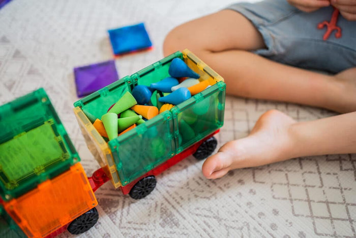 Toy vehicles built from colorful translucent magnetic tiles and wheels, filled with small cones on a patterned rug.