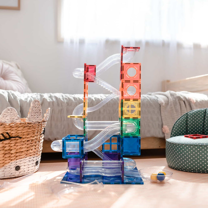 Tall rainbow magnetic tile ball run tower with clear slides and tubes on a light play mat in a sunny bedroom.