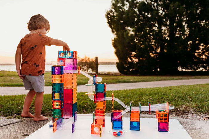 Child building a ball run with colorful translucent magnetic tiles and clear tubes on a lawn at sunset.