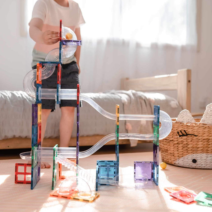 Child playing with a tall ball run made of colorful magnetic tiles and clear curved plastic tubes.