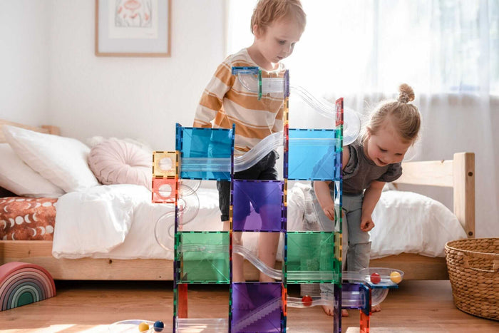 Two children play with a tall tower of colorful translucent magnetic tiles and clear plastic ball run tracks.
