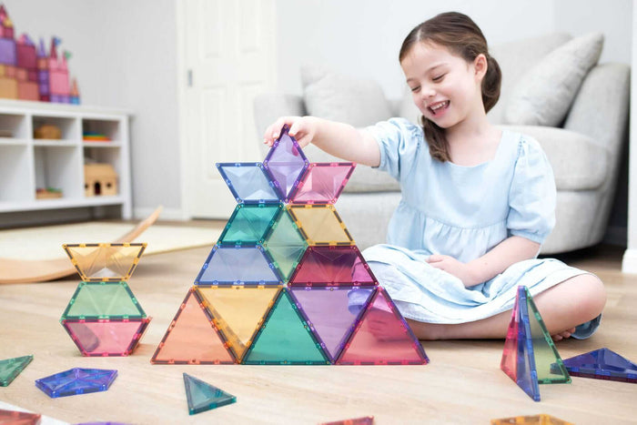 Girl builds a large structure using translucent pastel magnetic tiles in triangle, diamond, and hexagon shapes.