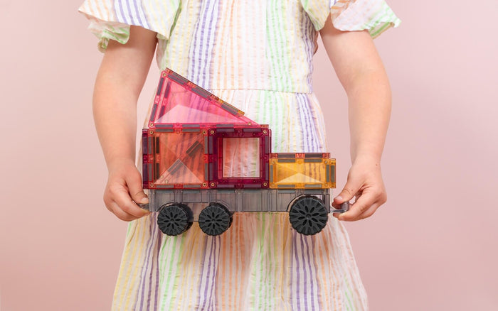 A child holds a toy vehicle built with translucent pink and orange magnetic tiles on a grey wheeled base.