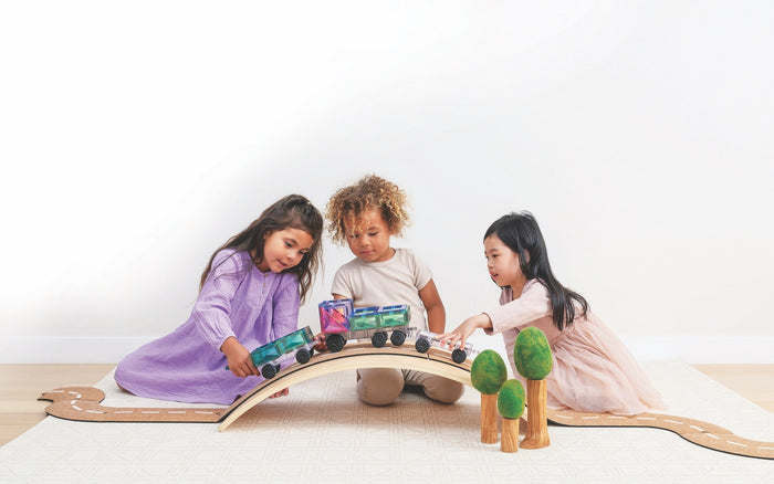 Three children play with pastel magnetic tile vehicles on a curved wooden bridge and cork road track.