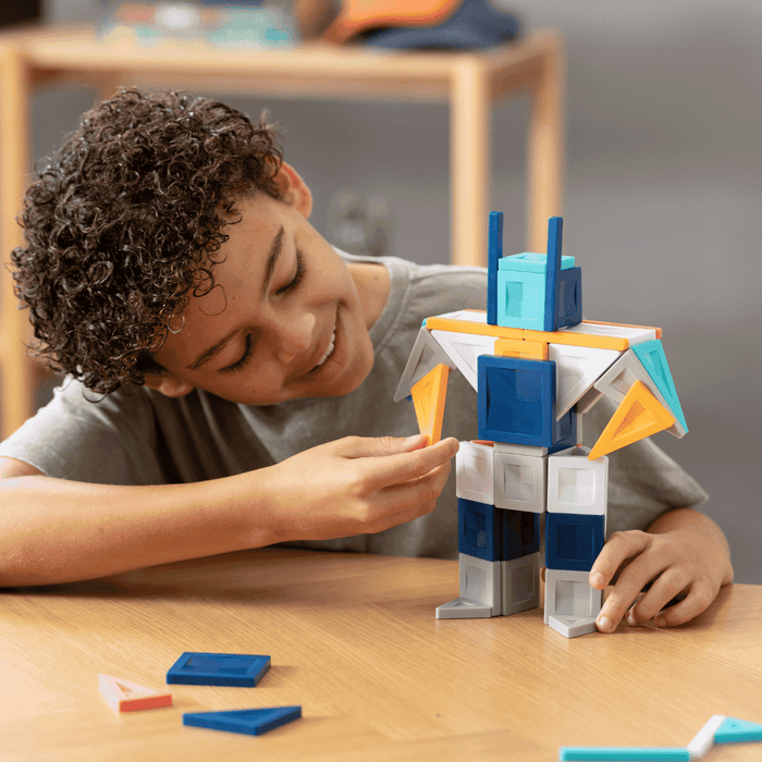 Child building a blue and white robot with orange accents using magnetic tiles and 3D blocks on a table.