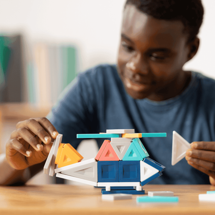 Boy building a toy helicopter using blue, orange, and red triangular and square magnetic tiles on a wooden table.