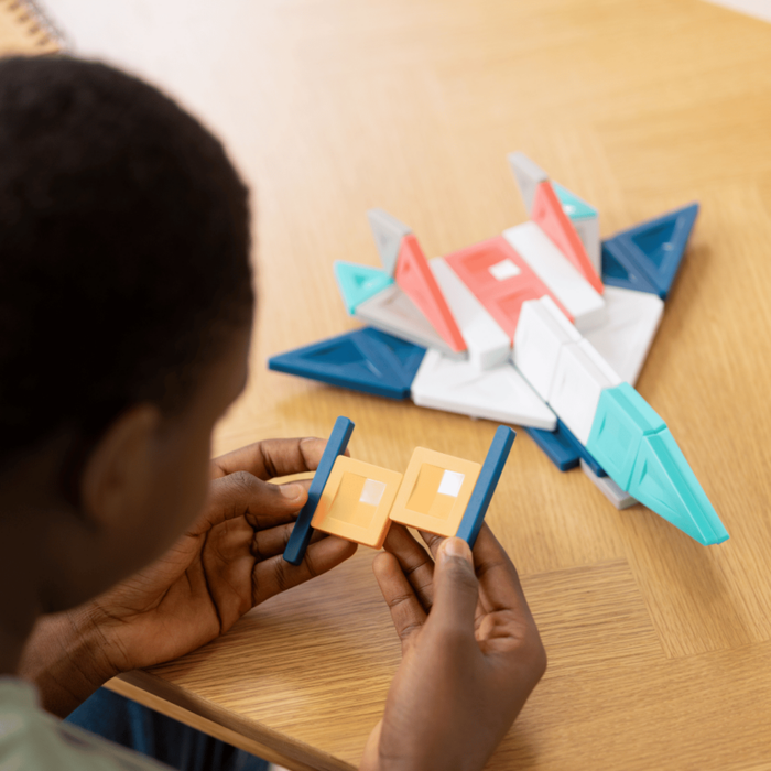 Child building a toy jet with colorful blue, red, and white geometric magnetic tiles on a wooden table.