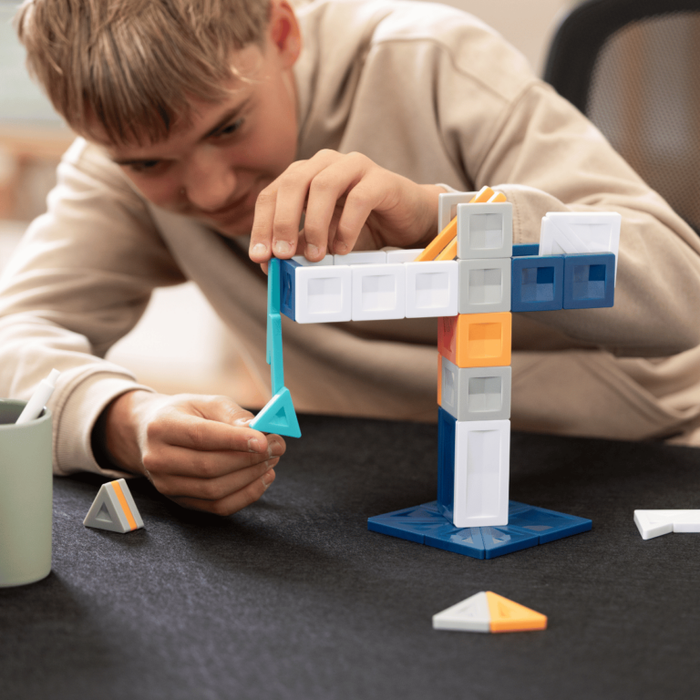 Teenager building a crane tower using white, blue, and orange magnetic tiles and rectangles on a dark surface.