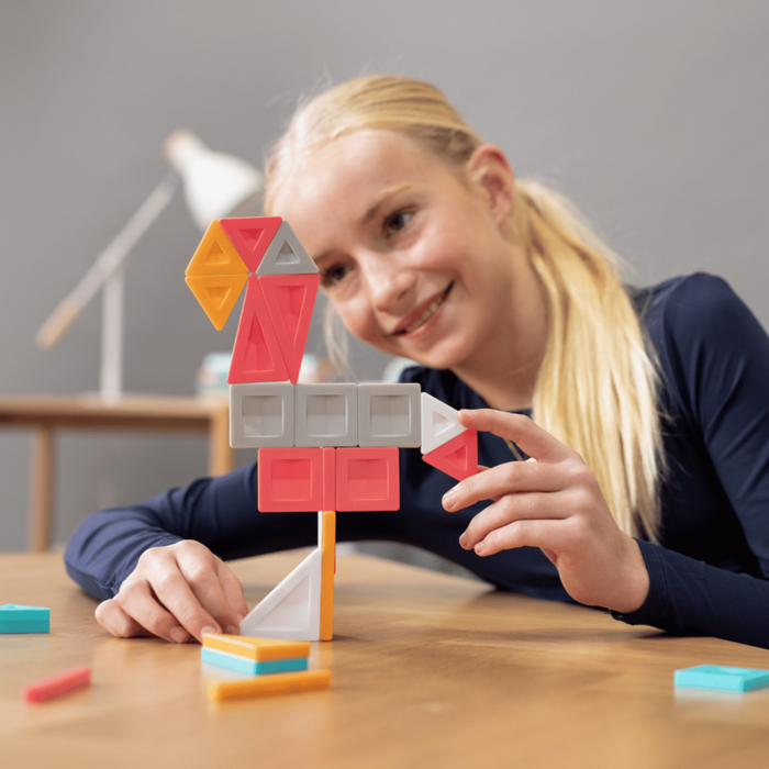 Smiling girl building a bird figure with red, orange, and grey magnetic tiles on a wooden table.