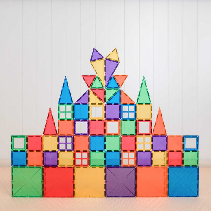 A symmetrical castle built from colorful rainbow magnetic square and triangle tiles against a white wood-paneled wall.