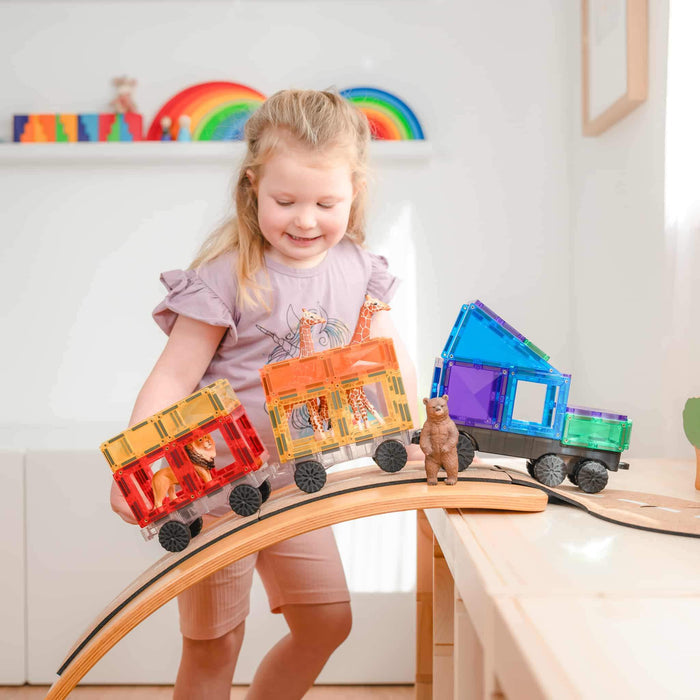 Child playing with a colorful magnetic tile train carrying toy animals down a wooden arched bridge.