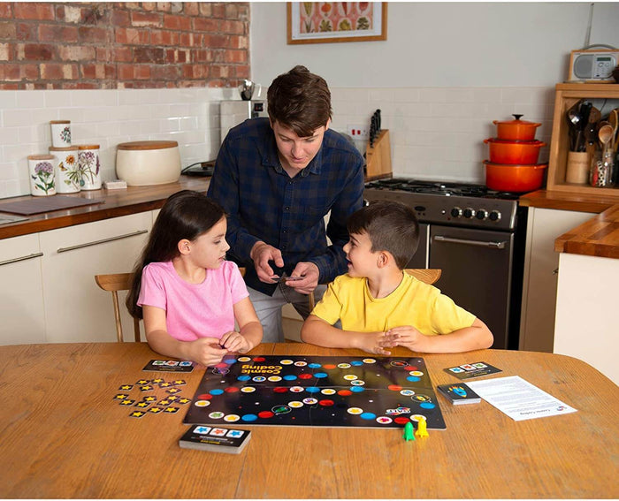 Two children and a man play a space-themed board game with colorful planet markers and star tokens on a wooden table.