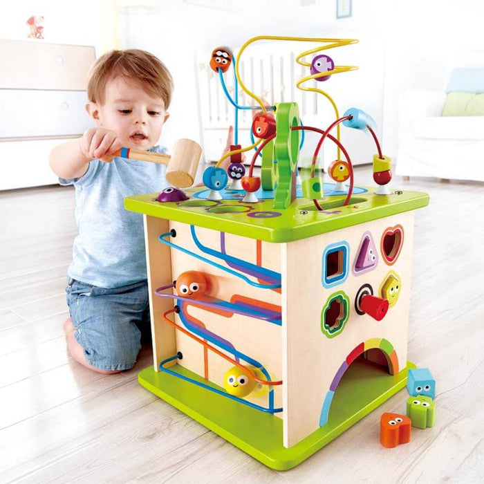 Toddler playing with a colorful wooden activity cube featuring a bead maze, animal slides, and a shape sorter.