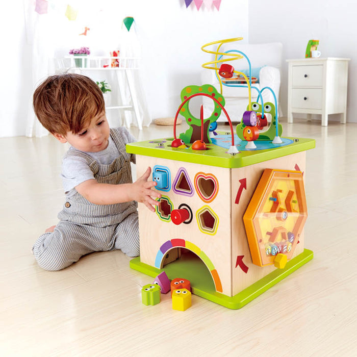 Toddler playing with a wooden activity cube featuring wire bead mazes and various colorful shape-sorting cutouts.