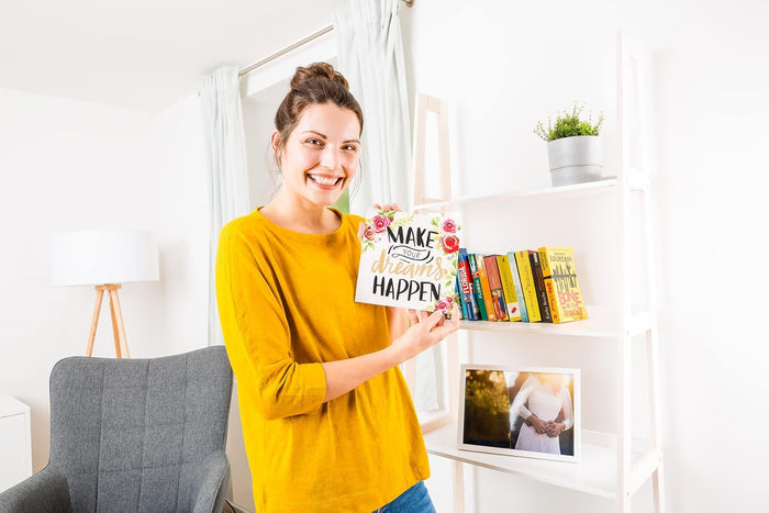 Smiling woman in yellow holds a floral "MAKE YOUR dreams HAPPEN" sign in a bright room with white shelves.