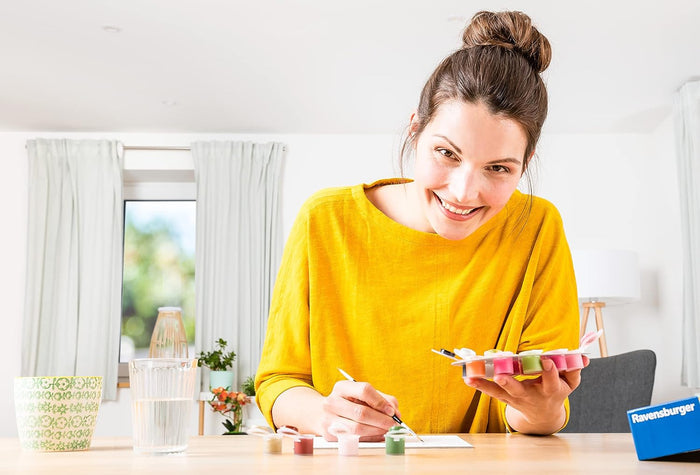 Smiling woman in a yellow shirt using a paint-by-numbers kit with small colorful paint pots at a table.