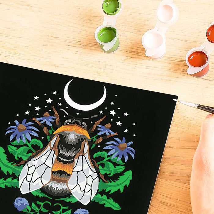 Paint-by-number kit of a bee under a crescent moon on a black canvas with paint pots and a brush.