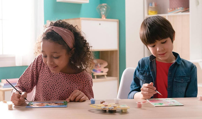 Two children sitting at a table painting detailed animal and floral canvases with small brushes and paint pots.