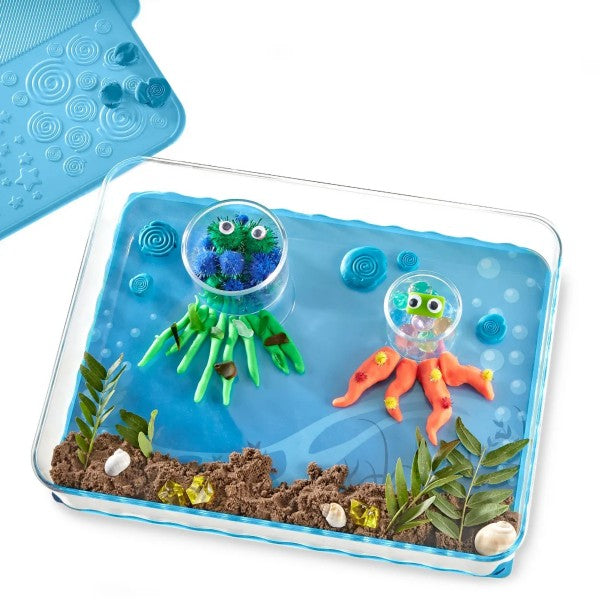Clear sensory tray with an underwater scene featuring clay jellyfish, sand, shells, and a blue textured mat.