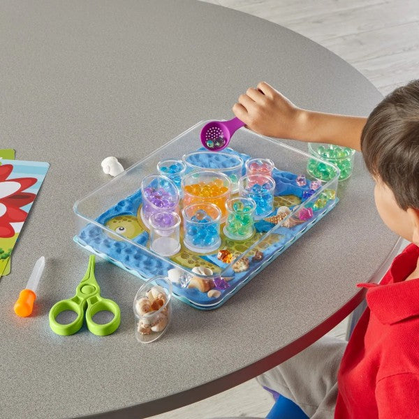 Child scoops colorful water beads into clear cups in a sensory tray with shells, a dropper, and plastic scissors.