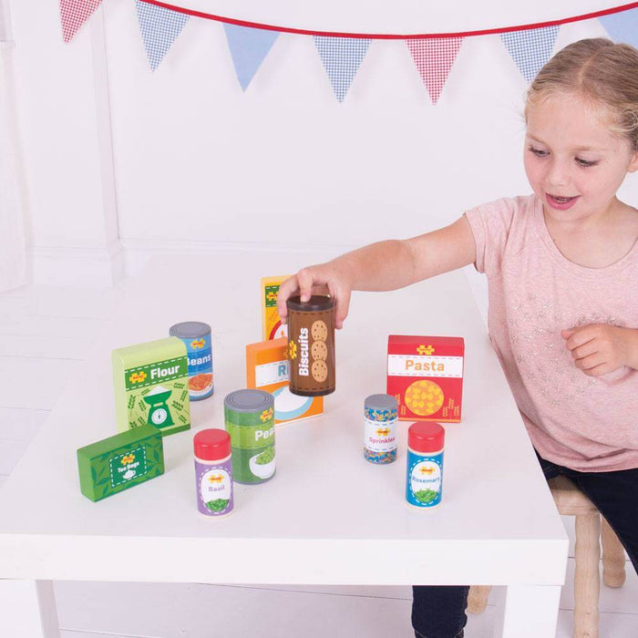 Young girl playing with colorful wooden toy grocery boxes and tins of pasta, flour, and biscuits on a white table.