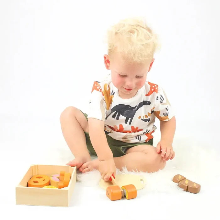 Toddler slices wooden bread on a board beside a crate of colorful toy pastries, including pretzels and rolls.