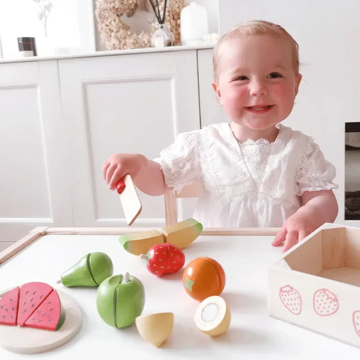 Smiling toddler playing with colorful wooden fruit slices and a toy knife on a white table with a storage crate.