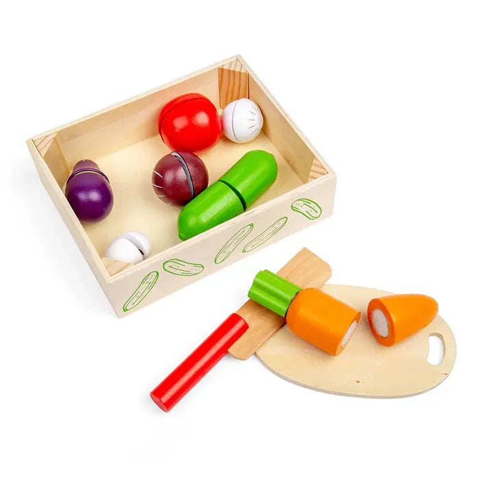 Wooden toy vegetables in a crate with a red-handled knife cutting an orange carrot on a small wooden board.