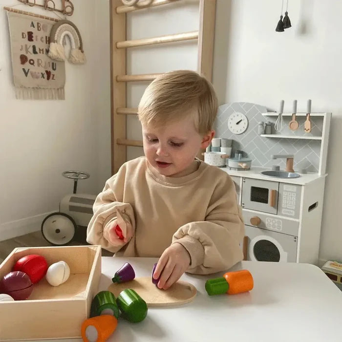 Toddler cutting colorful wooden play vegetables from a crate using a toy knife on a small wooden board.