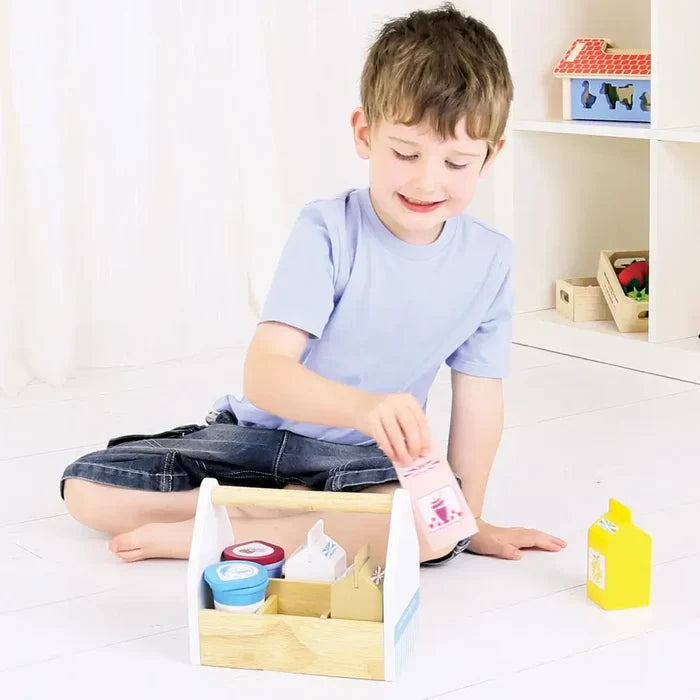 Child holding a pink toy milk carton next to a wooden crate with white, blue, and yellow dairy containers.