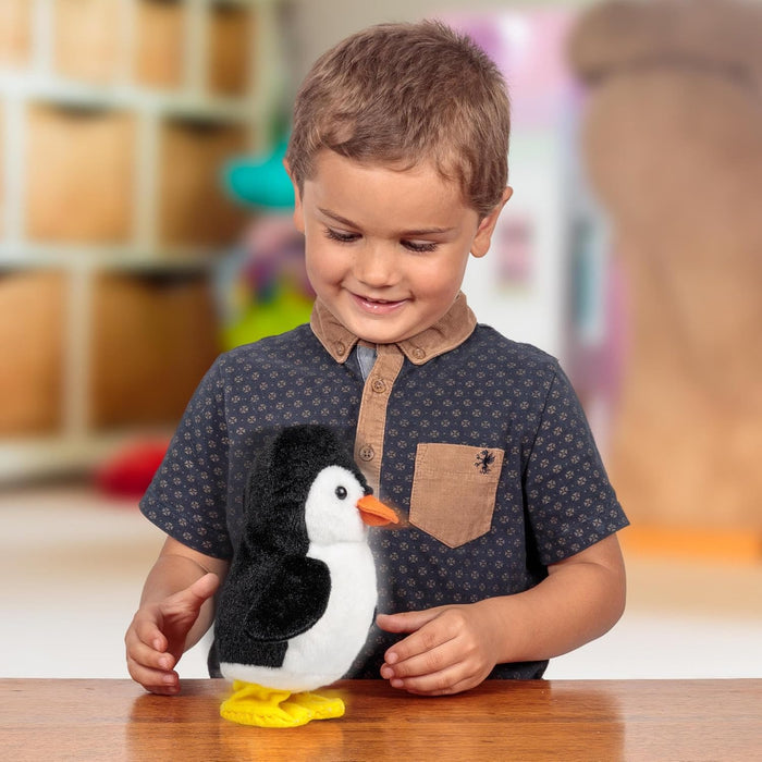 Smiling child looks at a black and white plush penguin with an orange beak and yellow feet on a wooden table.
