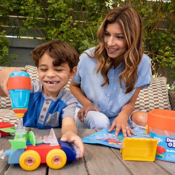 A boy builds a colorful toy car with a plastic drill as a woman watches, with an orange storage bucket nearby.