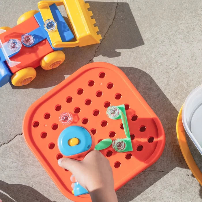 Child using a blue toy drill to screw clear bolts into an orange pegboard lid near a colorful toy bulldozer.