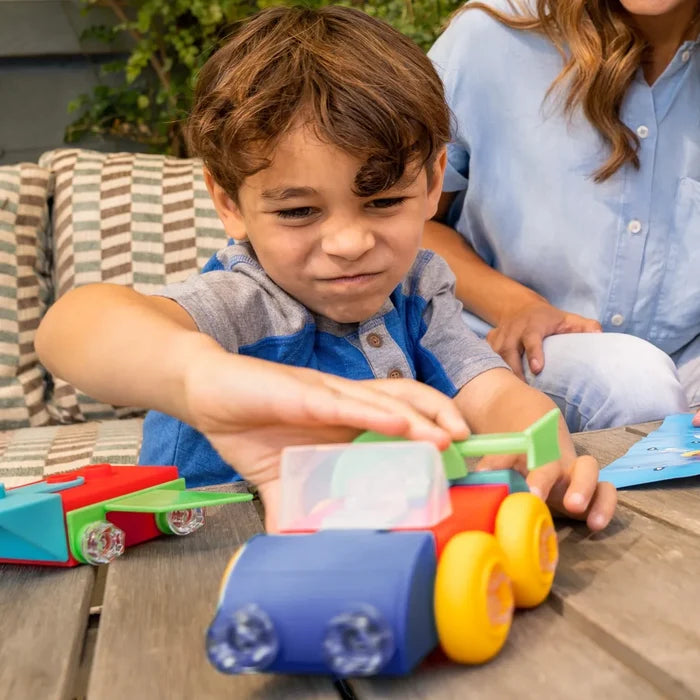 A boy builds colorful toy vehicles with chunky plastic parts and clear bolts on an outdoor wooden table.