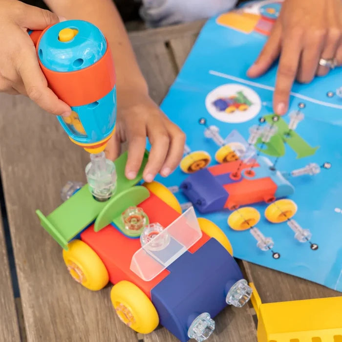 Child uses a toy power drill to build a colorful car with clear plastic bolts while following instructions.
