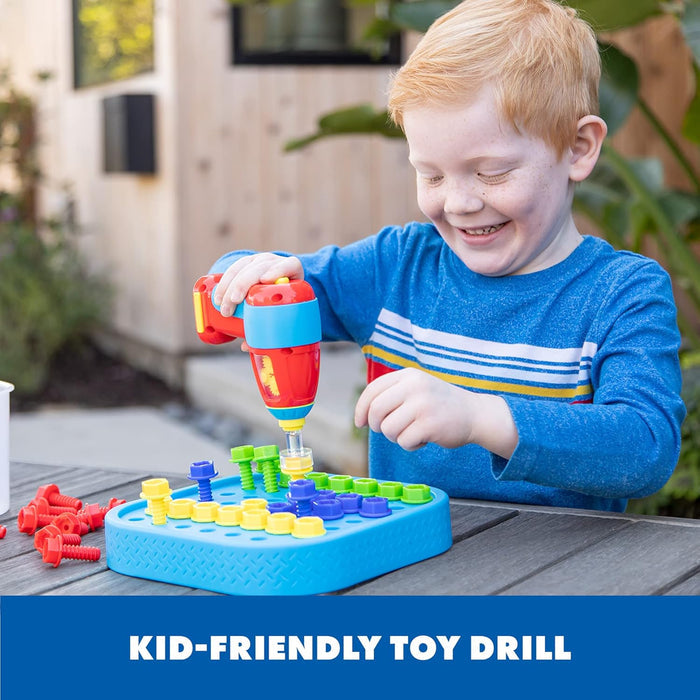Smiling boy uses a red toy drill to screw colorful plastic bolts into a blue activity board on a wooden table.