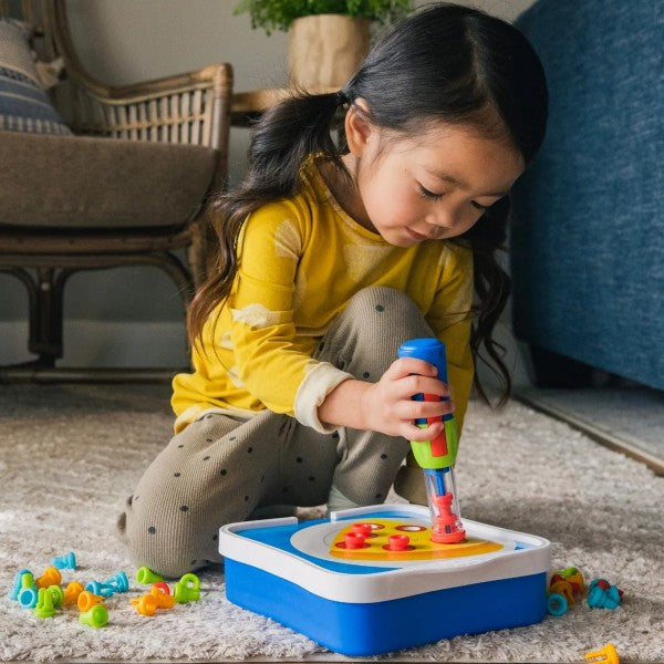 Child using a colorful toy tool to pop rivets into a blue activity tray with scattered plastic pieces on a rug.