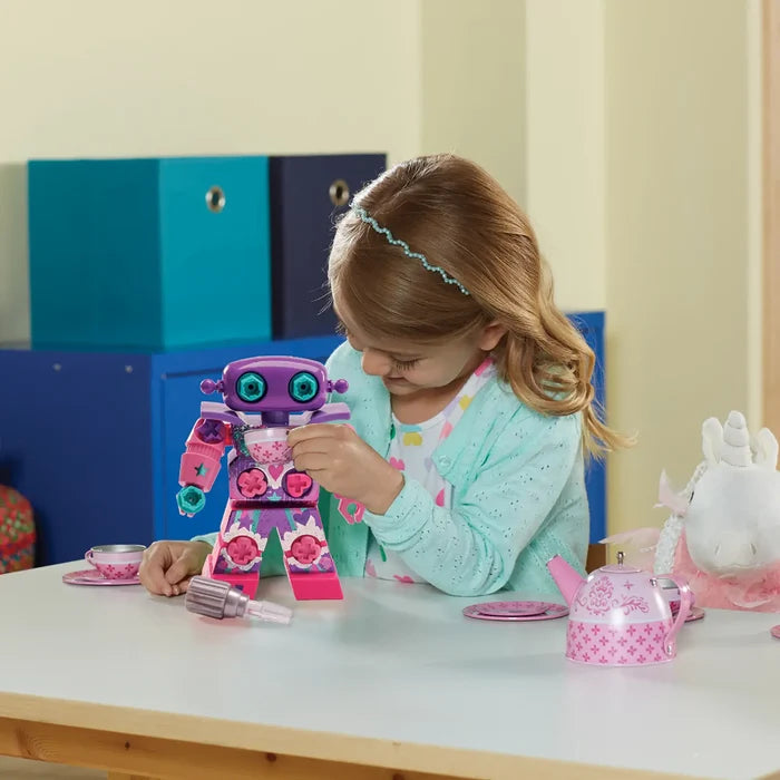 Girl using a toy drill on a purple and pink robot with teal eyes, sitting at a table with a pink tea set.