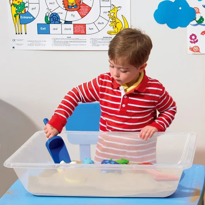 A child plays with sand and toys in a translucent, rectangular plastic tray resting on a blue tabletop.