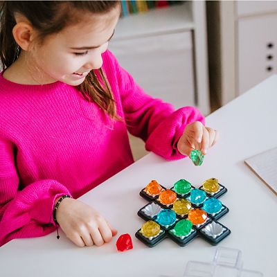 Child in pink sweater playing a logic puzzle with colorful translucent gem pieces on a black grid board.