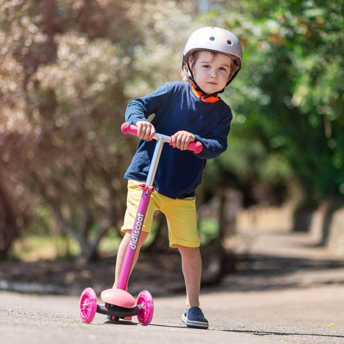 Child riding a pink 3-wheel kick scooter with pink wheels, pink grips, and an adjustable silver handle on a sunny path.