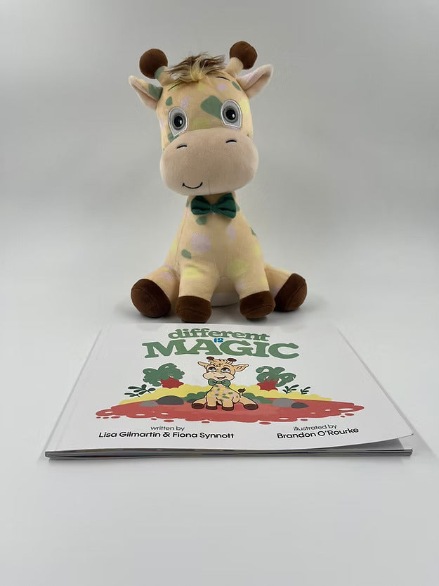 Plush yellow giraffe with pastel spots and green bow tie sitting behind the Different is Magic children's book.